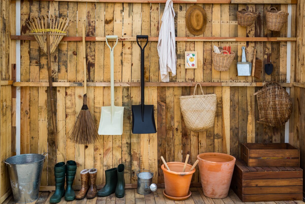 wooden shed wall containing a collection of some of the best gardening tools for beginners including baskets, rain boots, rakes, shovels, and a watering can