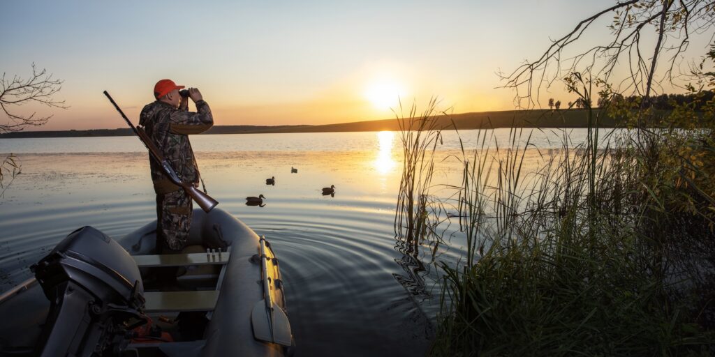 man looking out on the water with his binoculars while duck hunting from a boat near the shore