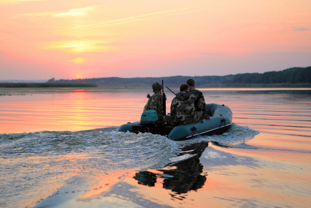 three men in camouflage riding on a boat through a lake on their way to a duck hunting mission at sunrise