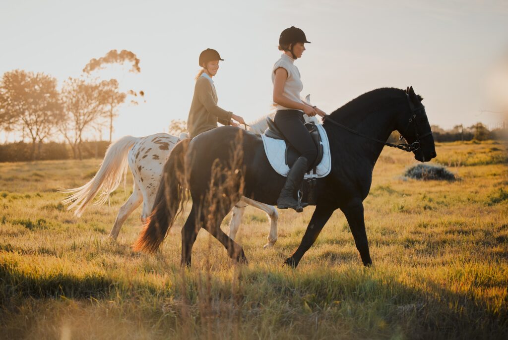 two women on horseback through a field at sunset wearing helmets as part of their horse riding equipment gear kit